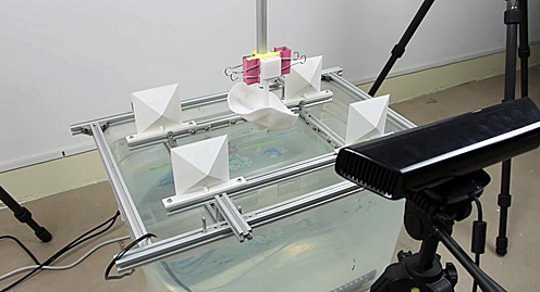 Setup includes a "gripper" mechanism (shown here holding a 3D mask), a water basin to hold the hydrographic color film, and a Kinect sensor to enable precise registration of the colors to the models surface.