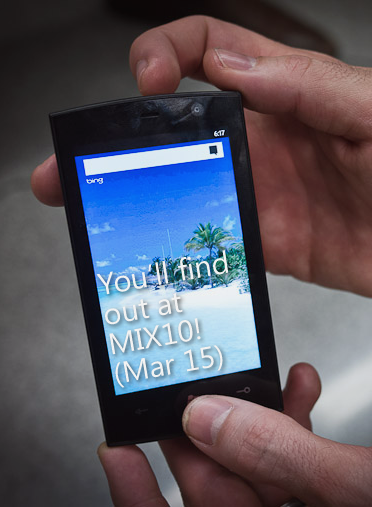 Hands holding Win 7 Phone that reads "You'll find out at MIX10! (Mar 15)"